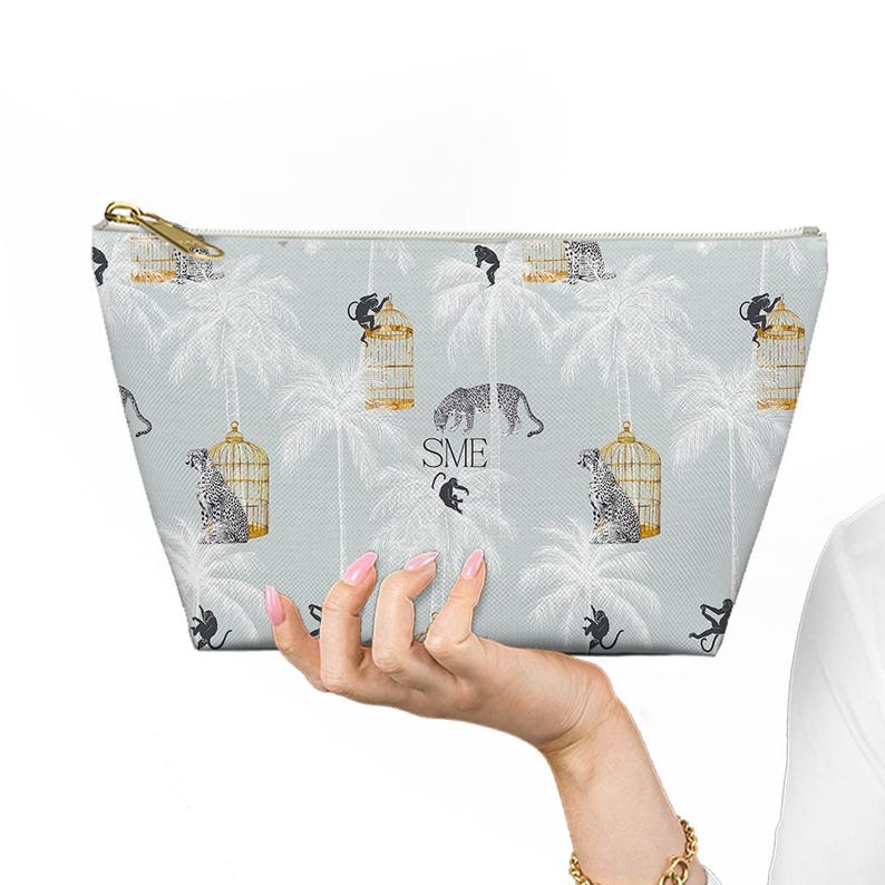 Peut inclure: Une trousse de maquillage bleu clair avec une fermeture &eacute;clair dor&eacute;e, pr&eacute;sentant un motif r&eacute;p&eacute;t&eacute; de palmiers, de singes et de l&eacute;opards. Le sac a un fond bleu clair avec les initiales "SME" imprim&eacute;es.