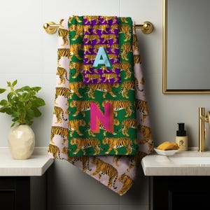 May include: A set of two decorative bath towels with a tiger print. One towel is green with orange and black tigers, and the other is pink with orange and black tigers. The letter "A" is on the green towel, and the letter "N" is on the green towel.