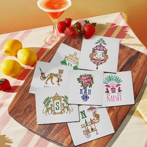 May include: A collection of white square coasters with various illustrations, including a leopard with a champagne bottle, monkeys, floral arrangements, and initial letters. Strawberries, lemons, and a cocktail glass are also present.