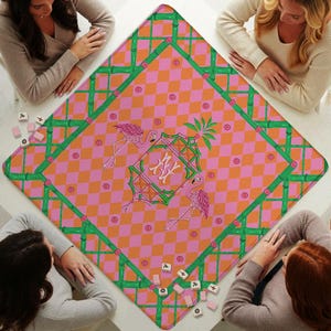 May include: A square game mat featuring a pink and orange checkered pattern with a green bamboo-style border. The center displays a decorative frame with the letters NY, flanked by two pink flamingos and small circular accents throughout.