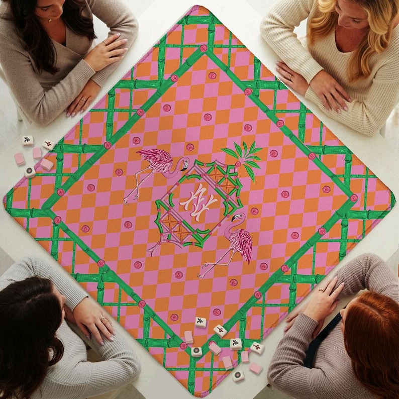 May include: A square game mat featuring a pink and orange checkered pattern with a green bamboo-style border. The center displays a decorative frame with the letters NY, flanked by two pink flamingos and small circular accents throughout.