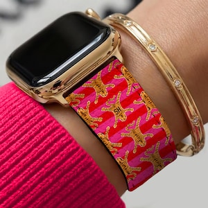 Könnte beinhalten: Goldene Apple Watch mit einem auffälligen Armband mit einem sich wiederholenden Tigermuster in Orange und Schwarz auf einem rosa-roten gestreiften Hintergrund. Ein goldenes Armband mit kleinen, klaren Steinen ist ebenfalls zu sehen.
