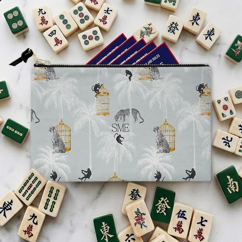 Peut inclure: Une pochette zipp&eacute;e bleu clair avec un motif tropical de palmiers, de l&eacute;opards et de singes. La pochette est entour&eacute;e de tuiles de Mahjong et de cartes de r&egrave;gles. Le texte "SME" est visible sur la pochette.