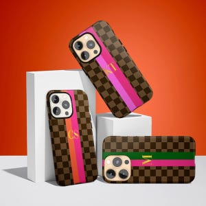 Puede incluir: Tres fundas de teléfono marrones con un patrón de cuadros y una franja rosa. Cada funda tiene una inicial diferente en dorado. Una funda está en posición vertical, otra en ángulo y otra plana.