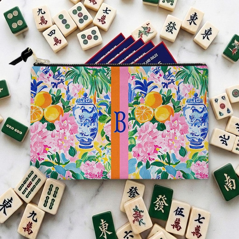 May include: A colorful zippered pouch with a floral design, oranges, and blue and white vases. The pouch has a vertical stripe with the letter "B". Mahjong tiles surround the pouch, with some visible game cards.