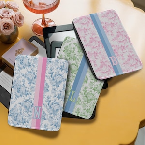 May include: Four floral-patterned Kindle covers in pink, blue, and green, each with a vertical stripe and a monogram letter. The covers are arranged on a yellow surface with a cocktail and a vase of roses in the background.