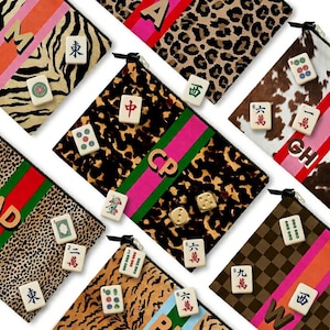 May include: An assortment of zippered pouches with various patterns, including leopard print, zebra stripes, and cowhide. Each pouch features a different design, with some displaying initials and others adorned with Mahjong tiles. The pouches are in various colors, including pink, green, and orange.