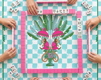 May include: A mahjong game set featuring a light blue and white checkered play mat with a central illustration of two monkeys holding pink umbrellas above a green urn with the letter M. Pink mahjong tiles are arranged in a square formation.