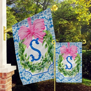 May include: A decorative garden flag featuring a pink bow and the letter "S" in blue, surrounded by a green wreath. The flag has a blue and white floral pattern border. A smaller matching flag is also visible.
