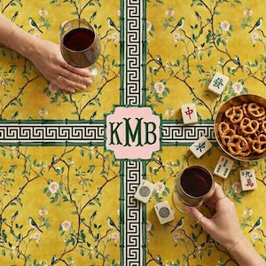 May include: A yellow tablecloth with a floral and bird pattern, green geometric borders, and a pink monogram. Wine glasses, pretzels, and Mahjong tiles are arranged on the table.