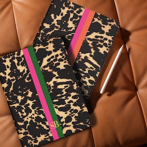 Op de afbeelding: Twee tablethoezen met een zwart en bruin dierenprintpatroon. Elke hoes heeft een verticale streep in roze, groen en oranje. Eén hoes heeft de gouden initialen "SB". Een witte stylus ligt op een bruin leren oppervlak.
