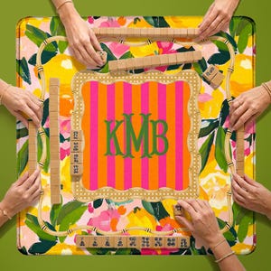 May include: A square board game mat featuring a vibrant lemon and floral pattern with a central orange and pink striped square containing the green monogram KMB. The mat is surrounded by wooden tile racks and tiles with black symbols.