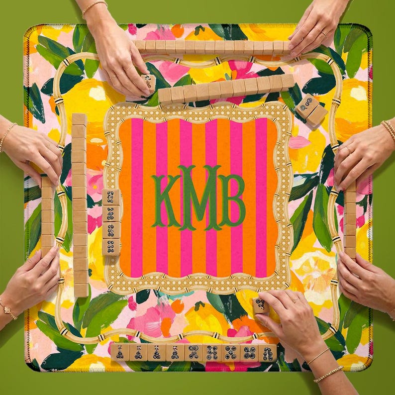 May include: A square board game mat featuring a vibrant lemon and floral pattern with a central orange and pink striped square containing the green monogram KMB. The mat is surrounded by wooden tile racks and tiles with black symbols.