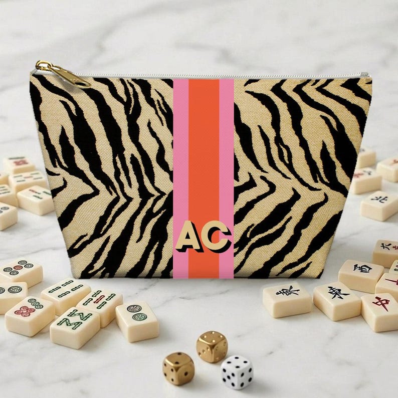 May include: A beige cosmetic bag with a black tiger stripe pattern and a vertical pink and orange stripe. The bag has gold-colored zipper and the initials "AC" in gold. Mahjong tiles and dice are scattered around the bag.