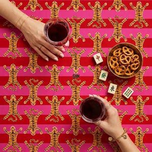 May include: A vibrant red and pink striped tablecloth with a repeating tiger pattern. Two glasses of red wine, a small bowl of pretzels, and Mahjong tiles are on the table. The letters "SHN" are visible on the tablecloth.