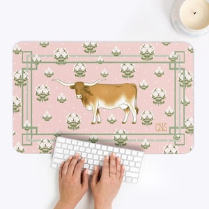 May include: A rectangular desk mat with a pink background featuring a brown longhorn illustration. The mat has a green border and white floral pattern. The initials "CNS" are in the lower right corner.