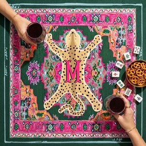 May include: A square, green and pink patterned game mat with a cheetah design and the letter "M". The mat is surrounded by game pieces, a bowl of pretzels, and two glasses of red wine.