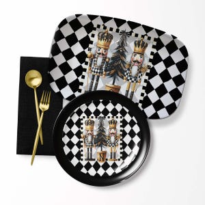 May include: A festive holiday table setting featuring a black and white checkered serving tray and plate. The tray and plate have a central image of nutcrackers and a Christmas tree. Gold utensils and a black napkin complete the setting.