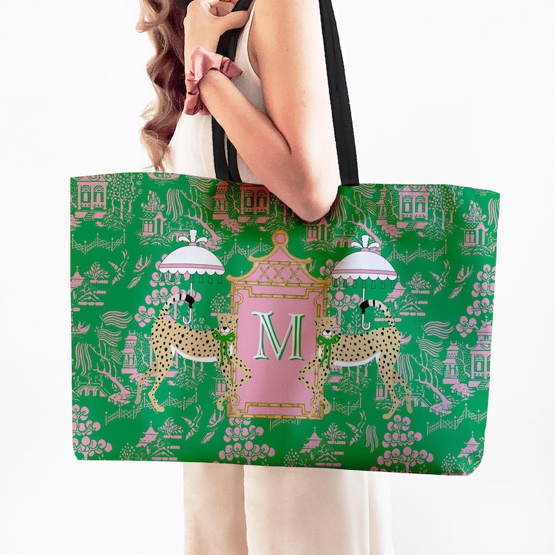 May include: Green tote bag with a pink and white Asian-inspired pattern. The bag features a pink frame with the letter "M" and two cheetahs holding umbrellas. Black straps complete the design.