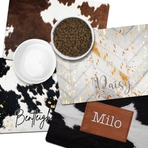 May include: Three cowhide dog bowls with personalized names:  "Bentleigh", "Daisy", and "Milo". The bowls are white ceramic with a brown and white cowhide pattern.