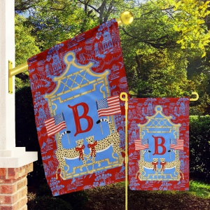 May include: A red and blue garden flag with a blue background and a gold bamboo frame. The frame encloses a blue square with the letter "B" in red. Two cheetahs with red bows are standing inside the frame. Two American flags are behind the cheetahs. The flag has a blue and white pattern with small houses and trees.