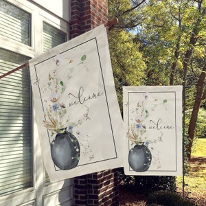 May include: Two welcome garden flags with a watercolor design of a vase with flowers. The flags are beige with black text that says "welcome".