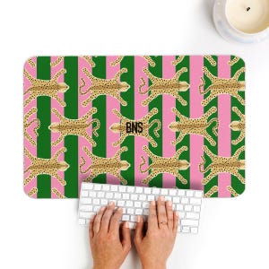 May include: A rectangular desk mat with a pink and green striped background. The mat features a repeating pattern of tan cheetahs with black spots. The initials "BNS" are in the center of the mat. A white keyboard is in front of the mat.