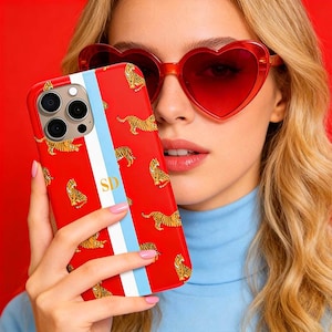 May include: Red phone case with a tiger print, a vertical blue and white stripe, and the initials "SD". The case is held in front of a person wearing heart-shaped red sunglasses and a blue turtleneck.