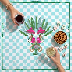 May include: A square, light blue and white checkered tablecloth with a green border. The tablecloth features a central design of a stylized palm tree with pink umbrellas and figures. A bowl of pretzels, mahjong tiles, and two glasses of wine are also present.