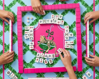 May include: A Mahjong game set featuring bright pink tiles and a central pink play mat decorated with a green leopard illustration and the letters SCF. The game board is surrounded by a light blue border with a green leafy vine pattern.