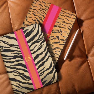 Op de afbeelding: Twee tablethoezen met dierenprints. Eén heeft een zebrapatroon, de andere een tijgerprint. Beide hebben een roze en oranje verticale streep. Eén heeft de initialen "LS". Een witte stylus is zichtbaar.