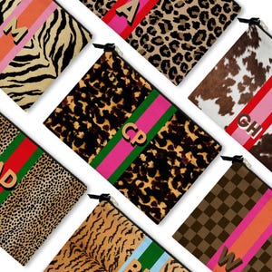 May include: A collection of zippered pouches with various animal print patterns, including zebra, leopard, and cowhide. Each pouch features a contrasting stripe and a letter or monogram in gold. The pouches are arranged on a white background.