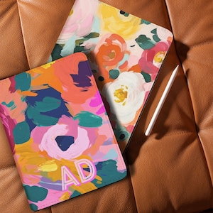 May include: Two colorful tablet cases with abstract floral designs in pink, orange, yellow, and teal. One case has the initials "AD" in a light pink font. A white stylus rests on the brown leather surface.