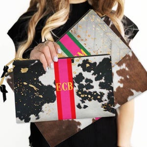 May include: A collection of stylish clutches in various designs. One clutch features a cowhide pattern with black and white spots, gold accents, and the initials "ECB" on a pink and red stripe. Another has a gray quilted pattern with gold flecks.