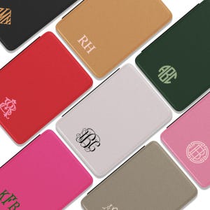 Puede incluir: Una colección de estuches rectangulares texturizados en varios colores, incluyendo negro, bronceado, rojo, verde oscuro, gris claro, rosa intenso y topo. Cada estuche presenta un monograma o iniciales únicas impresas en el frente.