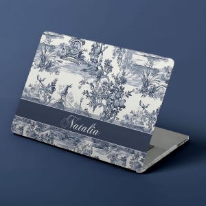 Peut inclure: Un ordinateur portable avec un motif de toile de Jouy bleu et blanc. Le design présente une scène de paysage avec des personnages, des arbres et des éléments architecturaux. Une bande bleu foncé en bas affiche le nom "Natalia" en écriture élégante.
