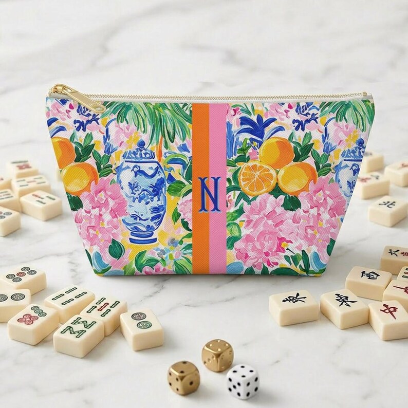 May include: A floral print cosmetic bag with a gold zipper, featuring oranges, pink flowers, and a blue and white vase design. The bag has a vertical stripe with the letter "N". Mahjong tiles and dice are scattered around the bag.
