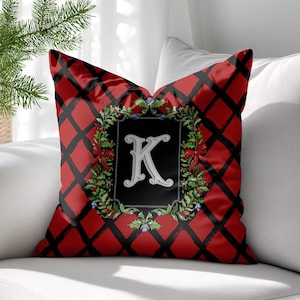 May include: Decorative square pillow with a red and black plaid pattern. The center features a black rectangle with a white letter "K" surrounded by a wreath of greenery, red bows, and blue berries. The pillow is on a white sofa.