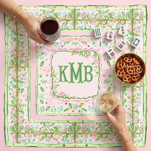 May include: A pink and green floral patterned square tablecloth with a monogram in the center. A gold bowl of pretzels, mahjong tiles, and glasses of wine are on the tablecloth.