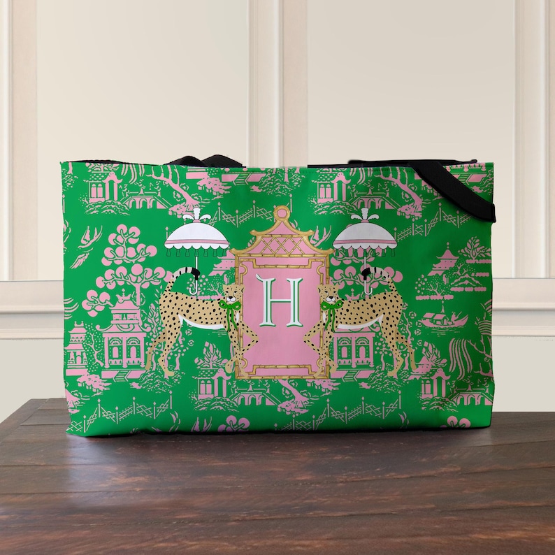 May include: Green tote bag with a pink and white Asian-inspired pattern. The design includes pagodas, flowers, and cheetahs. A pink frame with the letter "H" is centered on the bag. Black handles are visible.