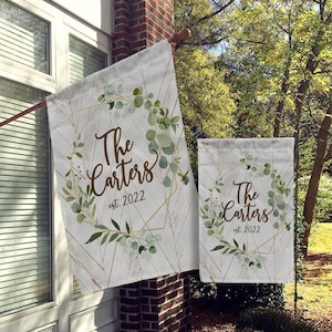 May include: Two white garden flags with a gold geometric design and green eucalyptus leaves. The text on the flags reads "The Carters est. 2022".