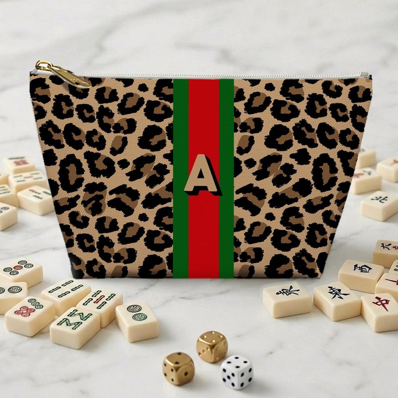 May include: A leopard print cosmetic bag with a gold zipper. The bag features a red and green stripe down the center with the letter "A" in the middle. Mahjong tiles and dice are scattered around the bag.