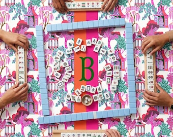 May include: A Mahjong game set featuring white tiles with black and red Chinese characters, arranged on a vibrant pink and orange patterned mat. The set includes four wooden tile racks, a central green letter B, and two small white dice.