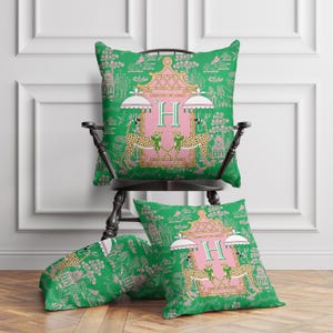 May include: Four decorative throw pillows with a green and pink Asian-inspired design. The pillows feature a pink pagoda with the letter "H", cheetahs, and parasols. The pillows are on a black chair and wooden floor.