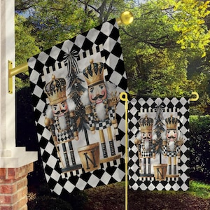 May include: Two black and white checkered garden flags with a design of three nutcracker figures. The nutcrackers are dressed in black and white checkered outfits with gold accents. The flags are hanging from poles in front of a green bush.
