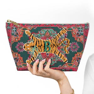 May include: A rectangular cosmetic bag with a colorful, patterned design. The bag features a tiger illustration with black stripes and the letter "B" in green. The bag has a gold zipper and a green, red, and orange floral pattern.