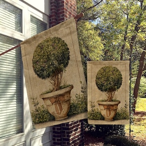 May include: Two burlap garden flags with a topiary design. The flags feature a green topiary tree in a brown pot with a bow on the front. The flags are hanging on a brick wall.