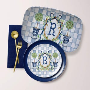 May include: A blue and white checkered serving tray and plate set with a decorative design. The design includes a monogrammed crest with the letter "R", topiary trees in blue vases, and a blue ribbon. Gold-colored spoon and fork are on a blue napkin.