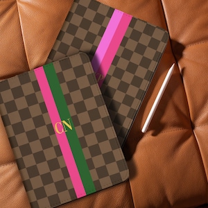 May include: Two brown and tan checkered tablet cases with a pink and green stripe. One case has the initials "CN" in gold. A white stylus rests on a brown leather surface.