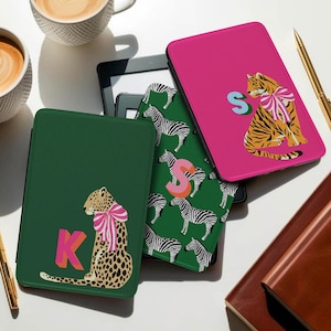 Könnte beinhalten: Eine Sammlung von Kindle-Hüllen in verschiedenen Farben und Designs. Eine Hülle ist dunkelgrün mit einem Leoparden mit rosa Schleife und dem Buchstaben "K". Eine andere ist pinkfarben mit einem Tiger mit rosa Schleife und dem Buchstaben "S". Eine dritte Hülle ist grün mit Zebrastreifen.